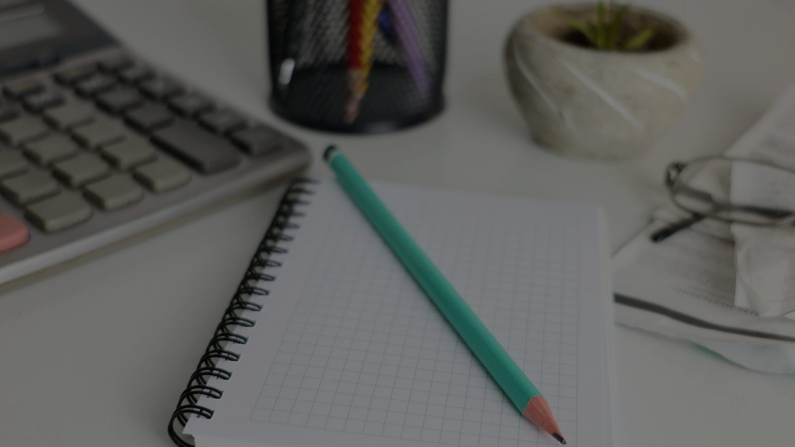 A calculator, a squared notebook with a green pencil and a small plant on a desk