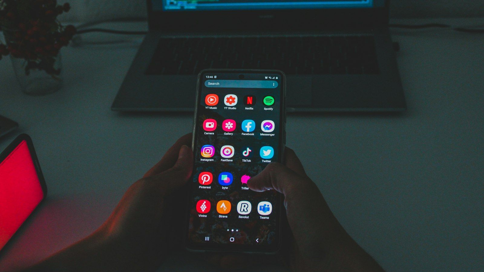 A hand holding a smartphone with a screen full of app icons, in the dark, in front of a laptop