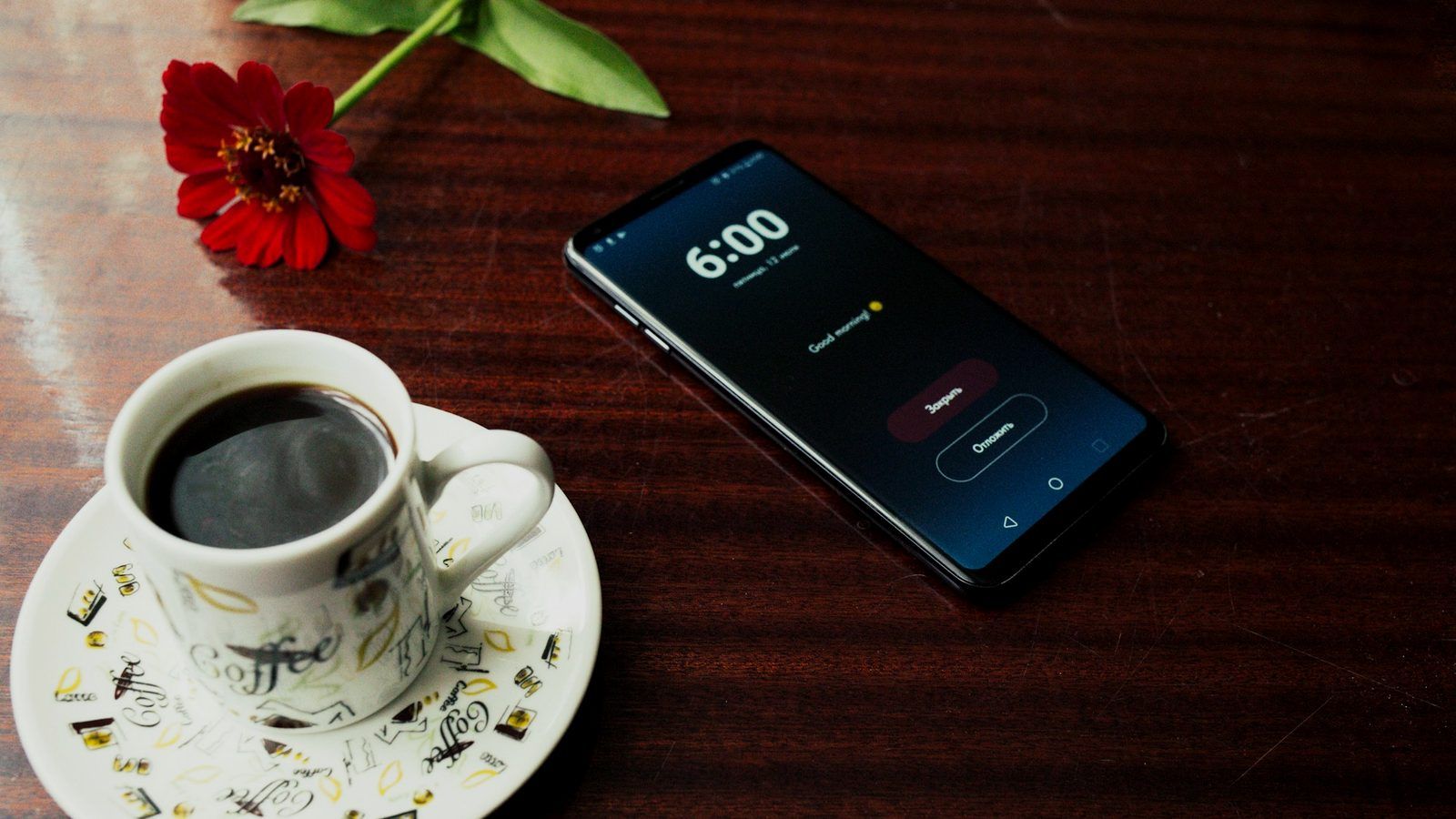 A smartphone and an espresso cup on a dark wooden table, with a red flower